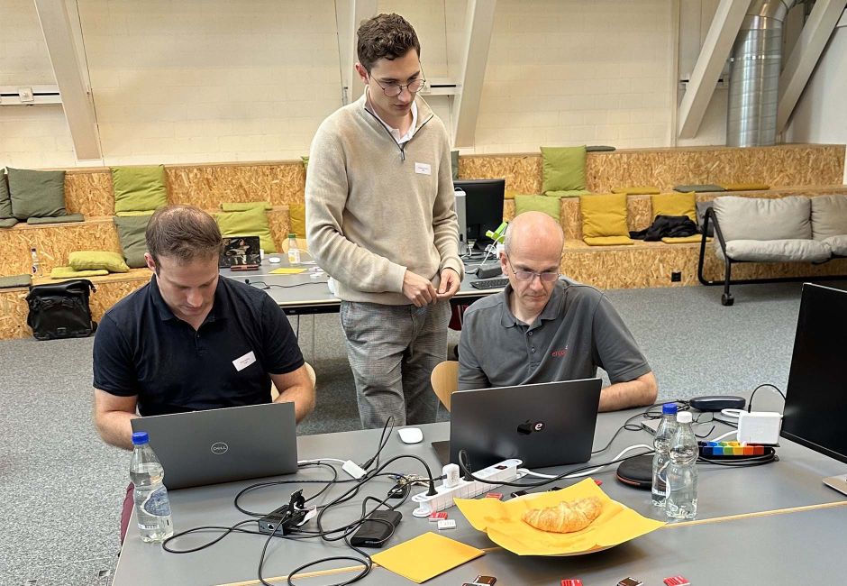 Zwei Engineers an einem Tisch am Laptop, 1 Person stehend dahinter am Beraten Zwei Engineers an einem Tisch am Laptop, 1 Person stehend dahinter am Beraten